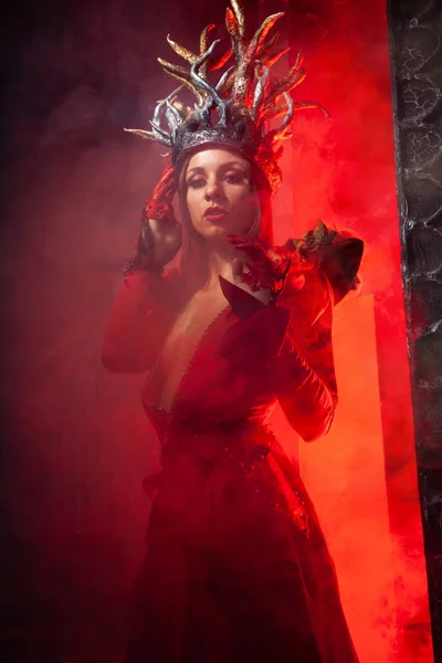 Bella donna strega di moda con le corna in forma di radici di albero in un lungo vestito di lusso sullo sfondo di un enorme cancello con fumo rosso. Concetto Halloween — Foto Stock