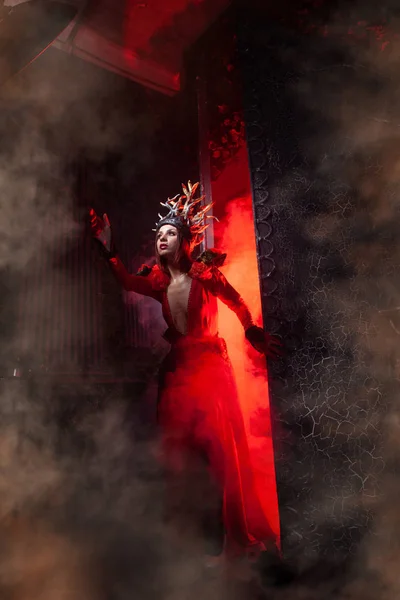 Hermosa mujer bruja de la moda con cuernos en forma de raíces de árbol en un vestido largo y lujoso en el fondo de una enorme puerta con humo rojo. Concepto Halloween — Foto de Stock