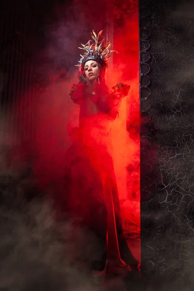 Beautiful fashion witch woman with horns in the form of tree roots in a long luxurious dress on the background of a huge gate with red smoke. Halloween concept — Stock Photo, Image