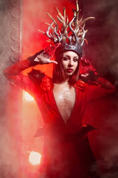Hermosa mujer bruja de la moda con cuernos en forma de raíces de árbol en un vestido largo y lujoso en el fondo de una enorme puerta con humo rojo. Concepto Halloween —  Fotos de Stock