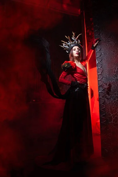 Schöne Mode Hexe Frau mit Hörnern in Form von Baumwurzeln in einem langen luxuriösen Kleid auf dem Hintergrund eines riesigen Tores mit rotem Rauch. Halloween-Konzept — Stockfoto