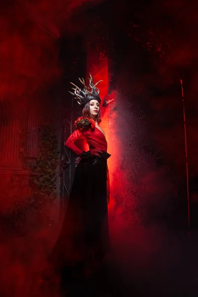 Hermosa mujer bruja de la moda con cuernos en forma de raíces de árbol en un vestido largo y lujoso en el fondo de una enorme puerta con humo rojo. Concepto Halloween — Foto de Stock