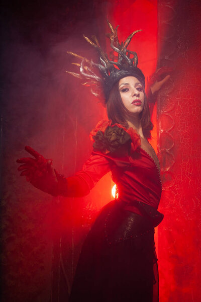 Beautiful fashion witch woman in a long luxurious dress on the background of a huge gate with red smoke. Halloween concept