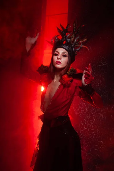 Beautiful fashion witch woman with horns in the form of tree roots in a long luxurious dress on the background of a huge gate with red smoke. Halloween concept — Stock Photo, Image