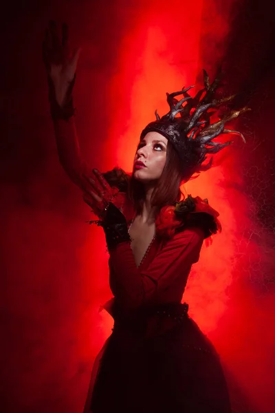 Beautiful fashion witch woman with horns in the form of tree roots in a long luxurious dress on the background of a huge gate with red smoke. Halloween concept — Stock Photo, Image