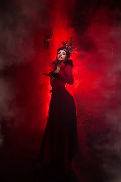 Beautiful fashion witch woman with horns in the form of tree roots in a long luxurious dress on the background of a huge gate with red smoke. Halloween concept — Stock Photo, Image