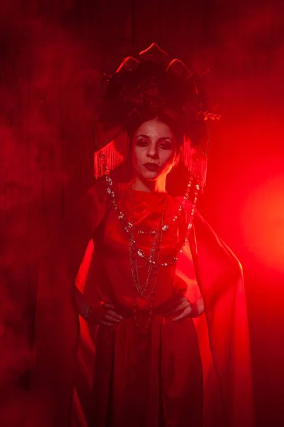 Chica con estilo estricto en un vestido rojo largo de moda y un sombrero voluminoso en el fondo de humo en el estudio por la noche solo. Concepto Halloween . — Foto de Stock