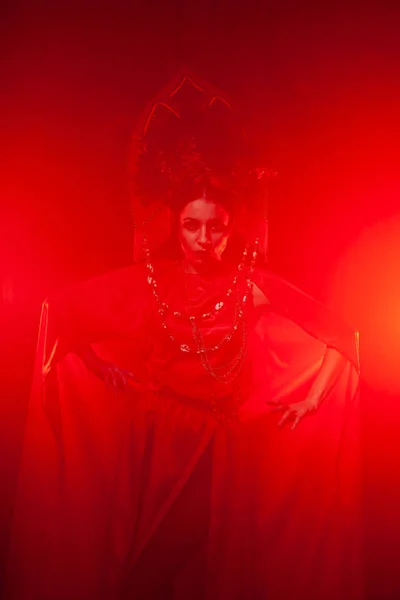 Chica con estilo estricto en un vestido rojo largo de moda y un sombrero voluminoso en el fondo de humo en el estudio por la noche solo. Concepto Halloween . —  Fotos de Stock