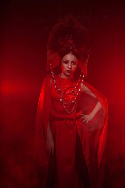 Menina elegante estrita em um vestido vermelho longo na moda e um chapéu volumoso no fundo da fumaça no estúdio à noite sozinho. Conceito de Halloween . — Fotografia de Stock
