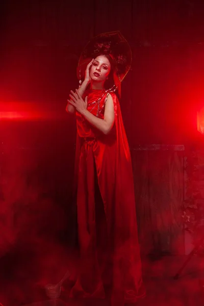 Ścisła stylowa dziewczyna w modnej długiej czerwonej sukni i obszerny kapelusz na tle dymu w studiu w nocy sam. Koncepcja Halloween. — Zdjęcie stockowe
