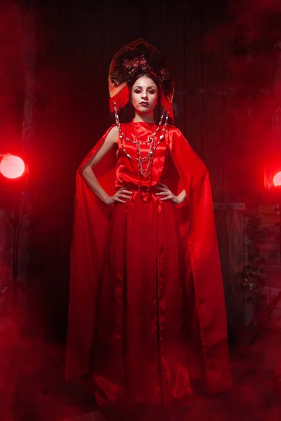 Menina elegante estrita em um vestido vermelho longo na moda e um chapéu volumoso no fundo da fumaça no estúdio à noite sozinho. Conceito de Halloween . — Fotografia de Stock