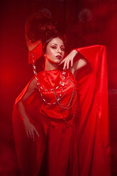 Rigorosa ragazza elegante in un abito rosso lungo alla moda e un cappello voluminoso sullo sfondo di fumo nello studio di notte da solo. Concetto Halloween . — Foto Stock