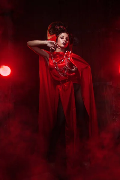 Das streng stylische Mädchen in einem modischen langen roten Kleid und einem voluminösen Hut vor dem Hintergrund des Rauchs allein in der Nacht im Studio. Halloween-Konzept. — Stockfoto