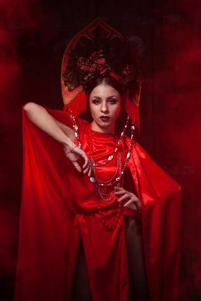 Strict stylish girl in a fashionable long red dress and a voluminous hat on the background of smoke in the Studio at night alone. Halloween concept. — Stock Photo, Image