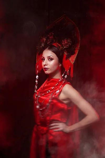 Rigorosa ragazza elegante in un abito rosso lungo alla moda e un cappello voluminoso sullo sfondo di fumo nello studio di notte da solo. Concetto Halloween . — Foto Stock