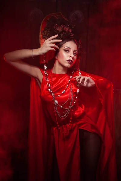 Rigorosa ragazza elegante in un abito rosso lungo alla moda e un cappello voluminoso sullo sfondo di fumo nello studio di notte da solo. Concetto Halloween . — Foto Stock