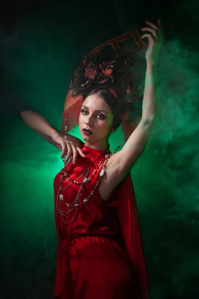 Ragazza rigorosa e spaventosa in un abito rosso lungo alla moda e un cappello voluminoso sullo sfondo di fumo verde nello Studio solo di notte. Concetto Halloween . — Foto Stock