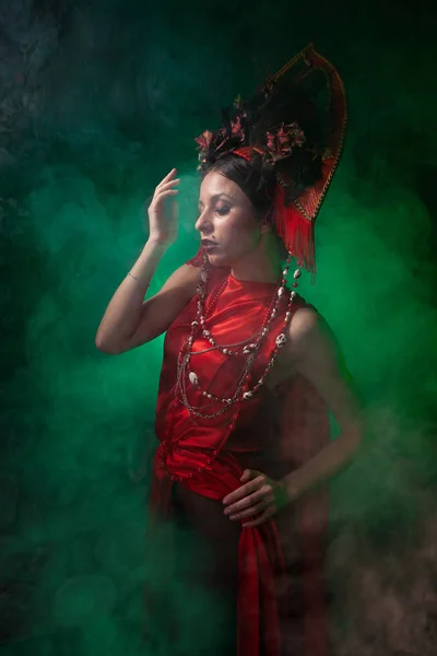 Ragazza rigorosa e spaventosa in un abito rosso lungo alla moda e un cappello voluminoso sullo sfondo di fumo verde nello Studio solo di notte. Concetto Halloween . — Foto Stock