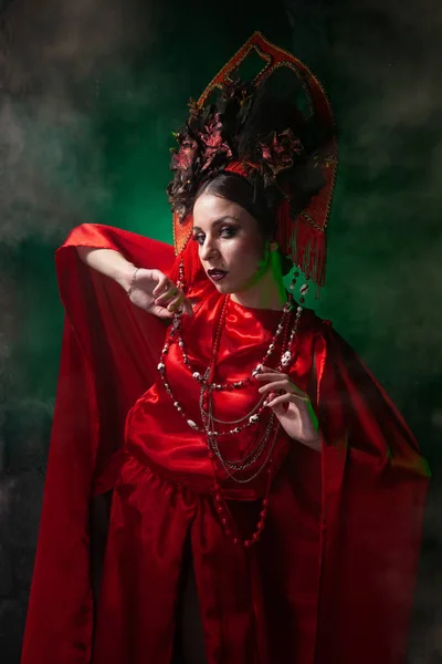 Strict and scary girl in a fashionable long red dress and a voluminous hat on the background of green smoke in the Studio at night alone. Halloween concept. — Stock Photo, Image