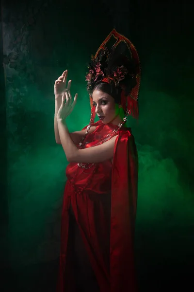 Ragazza rigorosa e spaventosa in un abito rosso lungo alla moda e un cappello voluminoso sullo sfondo di fumo verde nello Studio solo di notte. Concetto Halloween . — Foto Stock