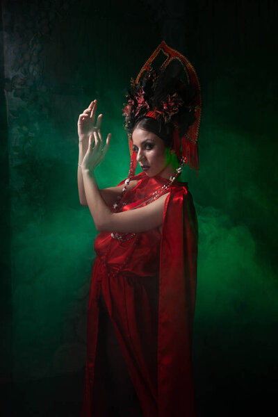 строгая и страшная девушка в модном длинном красном платье и объемной шляпе на фоне зеленого дыма в Студии ночью в одиночку. Концепция Хэллоуина
.