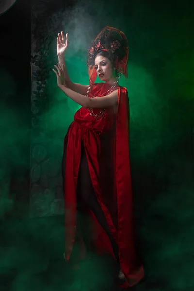Chica estricta y asustadiza en un vestido rojo largo de moda y un sombrero voluminoso en el fondo de humo verde en el estudio por la noche solo. Concepto Halloween . —  Fotos de Stock