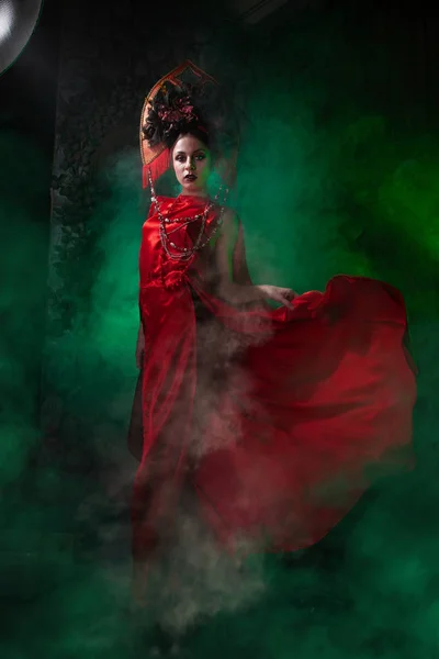 Ragazza rigorosa e spaventosa in un abito rosso lungo alla moda e un cappello voluminoso sullo sfondo di fumo verde nello Studio solo di notte. Concetto Halloween . — Foto Stock