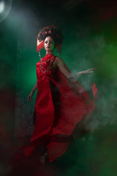 Strict and scary girl in a fashionable long red dress and a voluminous hat on the background of green smoke in the Studio at night alone. Halloween concept. — Stock Photo, Image