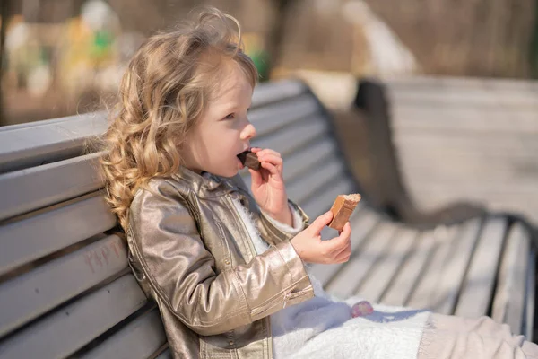 Emotioneel portret van een klein meisje met snoep, zittend op de Bank in het stadspark alleen buiten. mooi kind hebben een slechte smakelijke snack. — Stockfoto