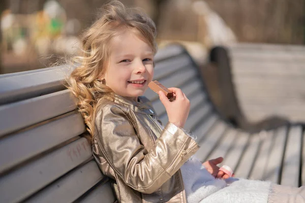 Emotional portrait of a little girl with sweets, sitting on the bench in the city park outdoor alone. pretty child have a bad tasty snack. — Stock Photo, Image