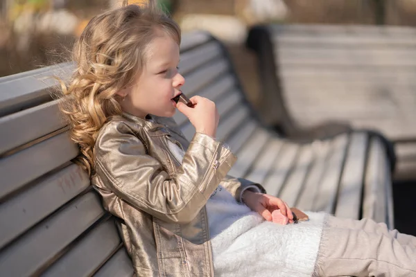Emotional portrait of a little girl with sweets, sitting on the bench in the city park outdoor alone. pretty child have a bad tasty snack. — Stock Photo, Image