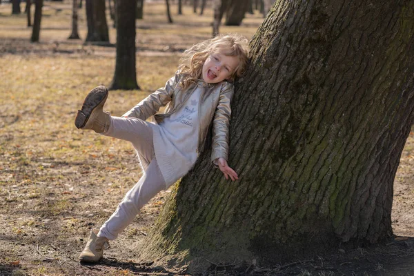 Jolie jeune fille bouclée en veste en cuir posant près de gros arbre dans le parc de la ville seul — Photo