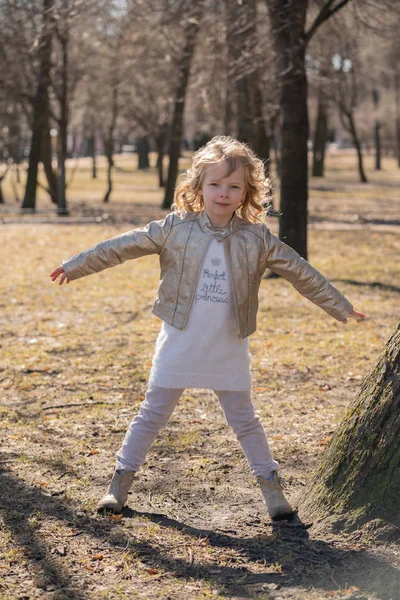 Pretty curly kid girl in leather jacket posing near big tree in the city park alone — Stock Photo, Image