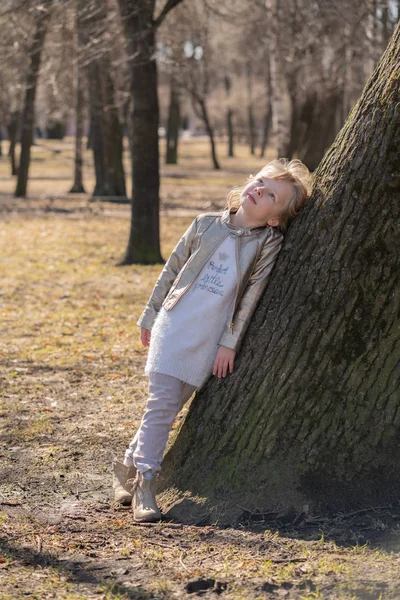 Muito encaracolado menina criança em jaqueta de couro posando perto de árvore grande no parque da cidade sozinho — Fotografia de Stock