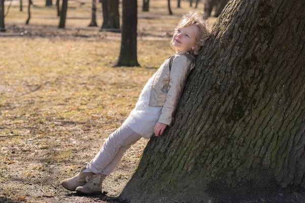 Muito encaracolado menina criança em jaqueta de couro posando perto de árvore grande no parque da cidade sozinho — Fotografia de Stock