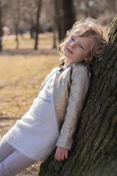 Ganska lockigt Kid flicka i skinnjacka poserar nära stora träd i stadsparken ensam — Stockfoto