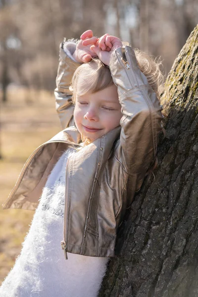 Ganska lockigt Kid flicka i skinnjacka poserar nära stora träd i stadsparken ensam — Stockfoto