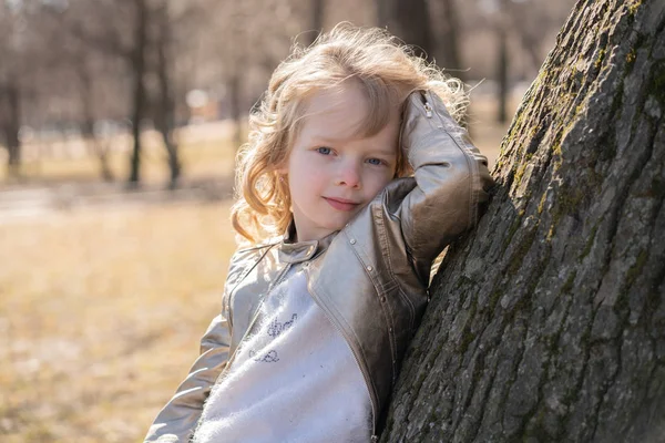 Ganska lockigt Kid flicka i skinnjacka poserar nära stora träd i stadsparken ensam — Stockfoto