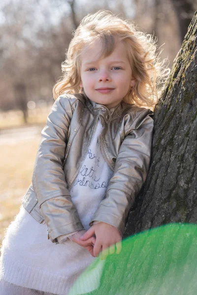 Pretty curly kid girl in leather jacket posing near big tree in the city park alone — Stock Photo, Image
