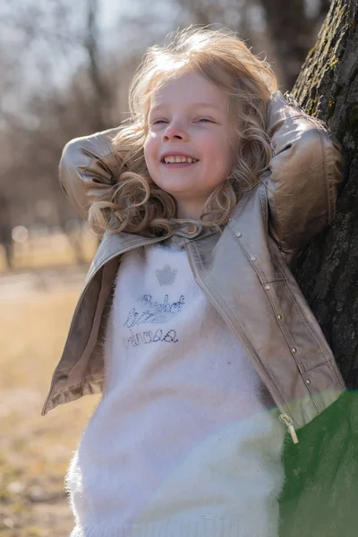 Pretty curly kid girl in leather jacket posing near big tree in the city park alone — Stock Photo, Image