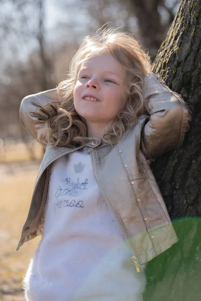 Pretty curly kid girl in leather jacket posing near big tree in the city park alone — Stock Photo, Image