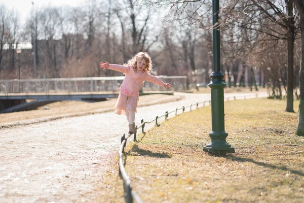 Pretty happy girl in pink jacket, cute skirt and warm pantyhose goes on curb on sunny day in the city park alone — Stock Photo, Image