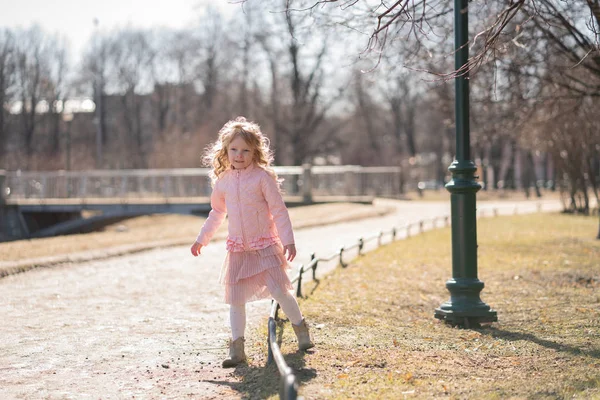 Ganska lycklig tjej i rosa jacka, Söt kjol och varma strumpbyxor går på trottoarkanten på solig dag i stadsparken ensam — Stockfoto