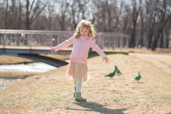 Jolie fille heureuse en veste rose, jupe mignonne et collants chauds court et effraie les oiseaux par une journée ensoleillée dans le parc de la ville seul — Photo