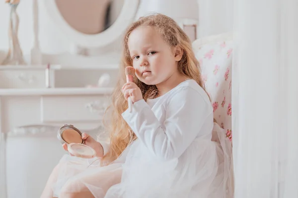 Portrét malé holčičky s make štětcem, který sedí na židli u velkého zrcadla v bílém pokoji — Stock fotografie