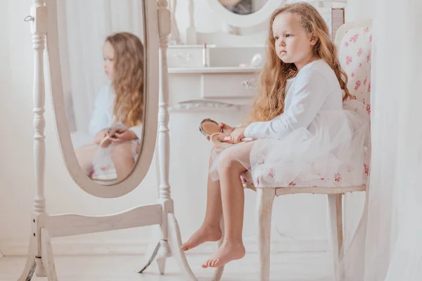 Closeup portrait of the little girl with make up brush sitting on the chair near the big mirror inside white room alone — Stock Photo, Image