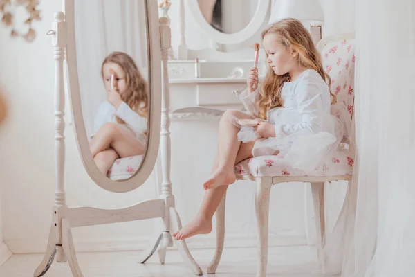 Портрет маленькой девочки с макияжем, сидящей на стуле рядом с большим зеркалом в белой комнате в одиночестве — стоковое фото