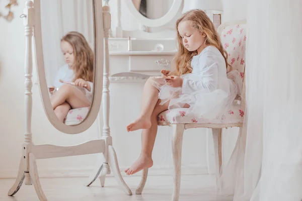 Closeup portrait of the little girl with make up brush sitting on the chair near the big mirror inside white room alone — Stock Photo, Image