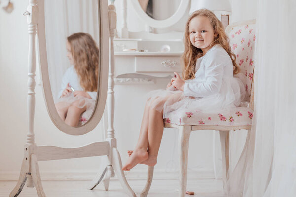 closeup portrait of the little girl with lipstick sitting on the chair near the big mirror inside white room alone