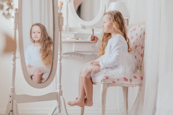 Closeup portrait of the little girl with lipstick sitting on the chair near the big mirror inside white room alone — Stock Photo, Image